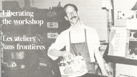A man with glasses and an apron holds a six-pack of bottles in a workshop.