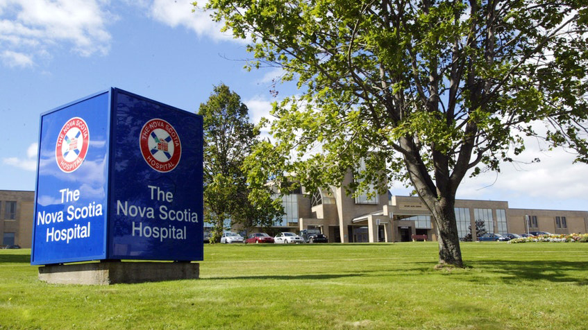 A blue hospital sign stands in front of a hospital building. There is a tree next to the sign.