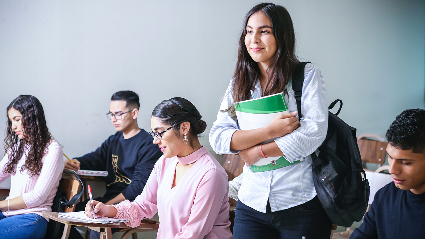 A young woman wearing a backpack stands holding a book in a classroom full of students.