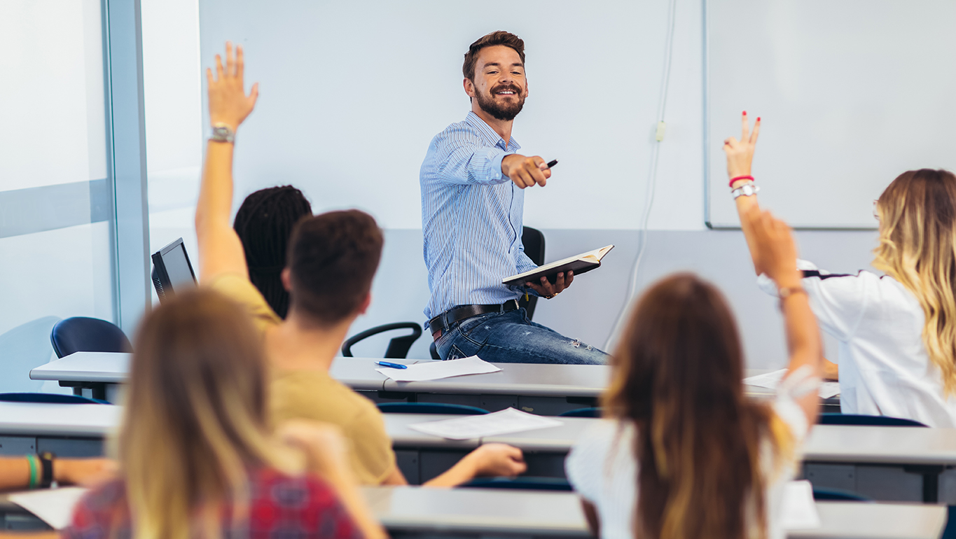 A teacher points to a student raising their hand in a classroom.